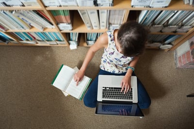 High angle view of a female student sitting against bookshelf with laptop on the library floor