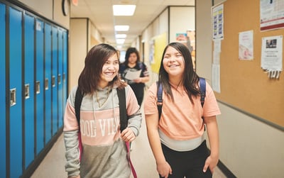 Two smiling female teen Canadian First Nations students walk in a school hallway between classes
